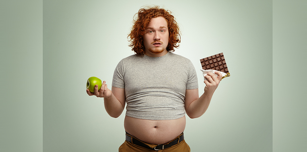 Indoor shot of uncertain confused plump young male facing hard choice as he has to chose between fresh organic apple in one hand and delicious bar of chocolate in other. Dilemma, diet and food
